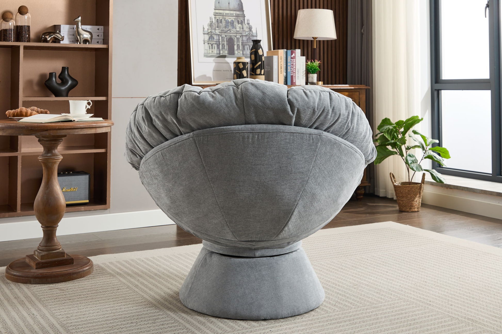 A modern living room showcases the Oversized Swivel Accent Chair— a large, gray, upholstered 360 swivel barrel chair—paired with a wooden side table, shelves, a potted plant, and natural light from the window.