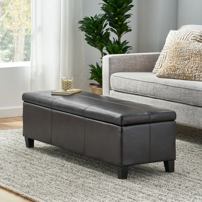 The Glouster - Contemporary Storage Ottoman, in dark brown leather, sits on a light rug before a gray sofa with patterned pillows and a green potted plant. A glass and book rest on the ottoman in this bright, modern living room.