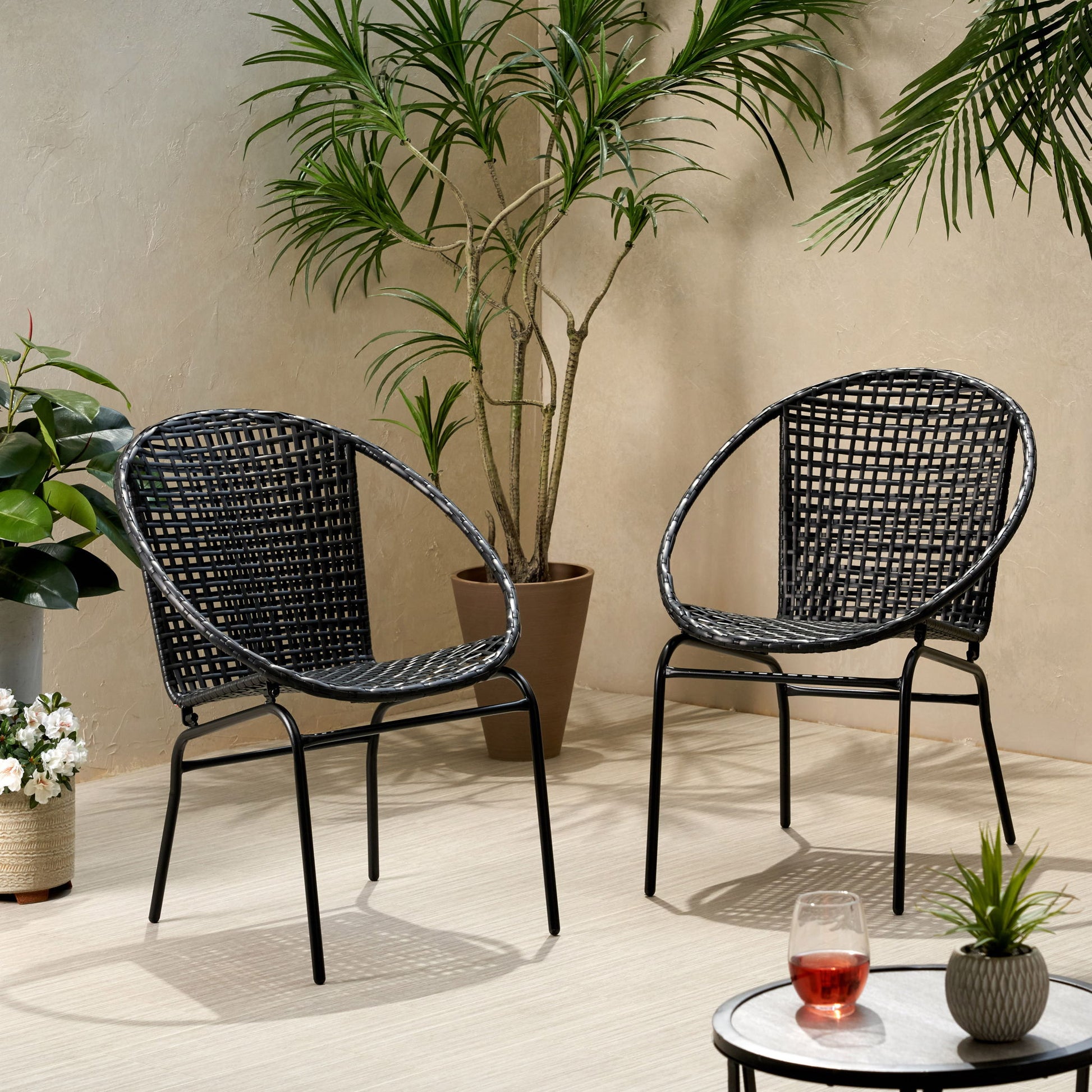 Two Java - Chair With Comfortable Design in black are set on a light patio floor among potted plants. In front, a small round table holds a glass of red drink and another glass, creating a calm and inviting setting.