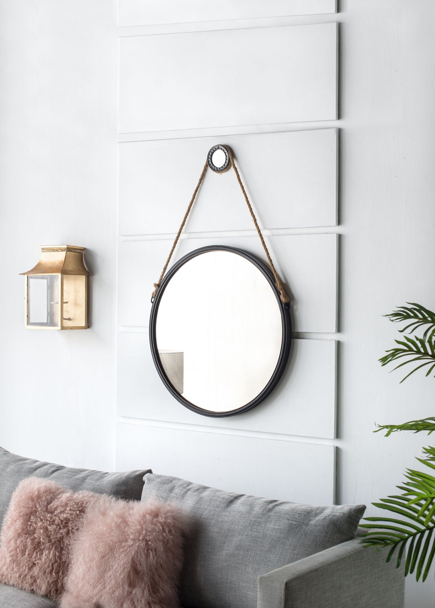 The Handsome Cleveland Mirror with Rope Strap, featuring a round metal frame, hangs on a white wall above a gray sofa with pink fuzzy pillows. A gold wall sconce and green plant accent the modern living room decor.