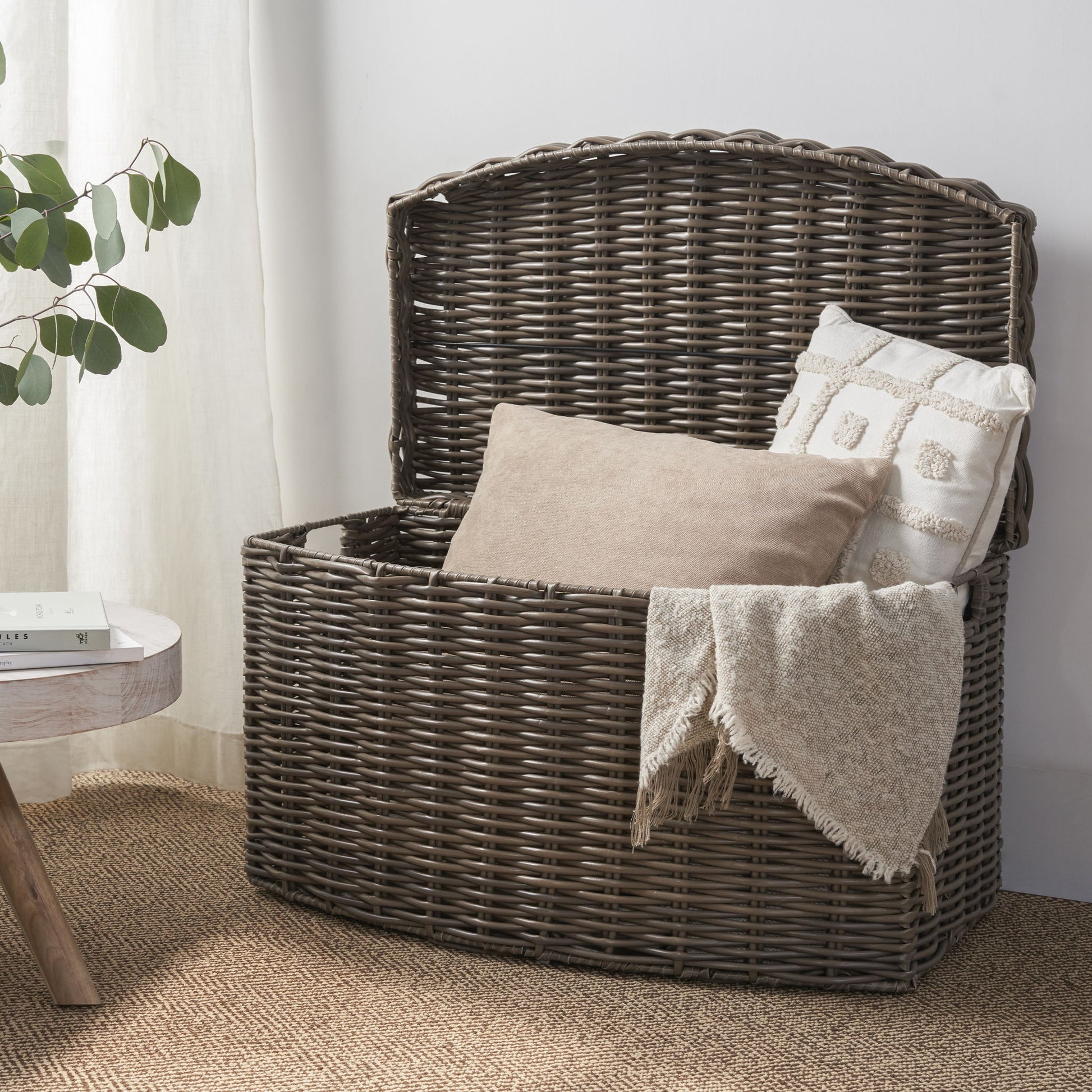 The Joseph Rectangular Curve Resin Woven Wicker Trunk in walnut brown, topped with a beige and white pillow and a fringed blanket, sits on a woven rug beside a small round table with a plant and book.