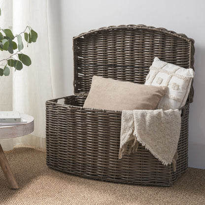 The Joseph Rectangular Curve Resin Woven Wicker Trunk in walnut brown, topped with a beige and white pillow and a fringed blanket, sits on a woven rug beside a small round table with a plant and book.