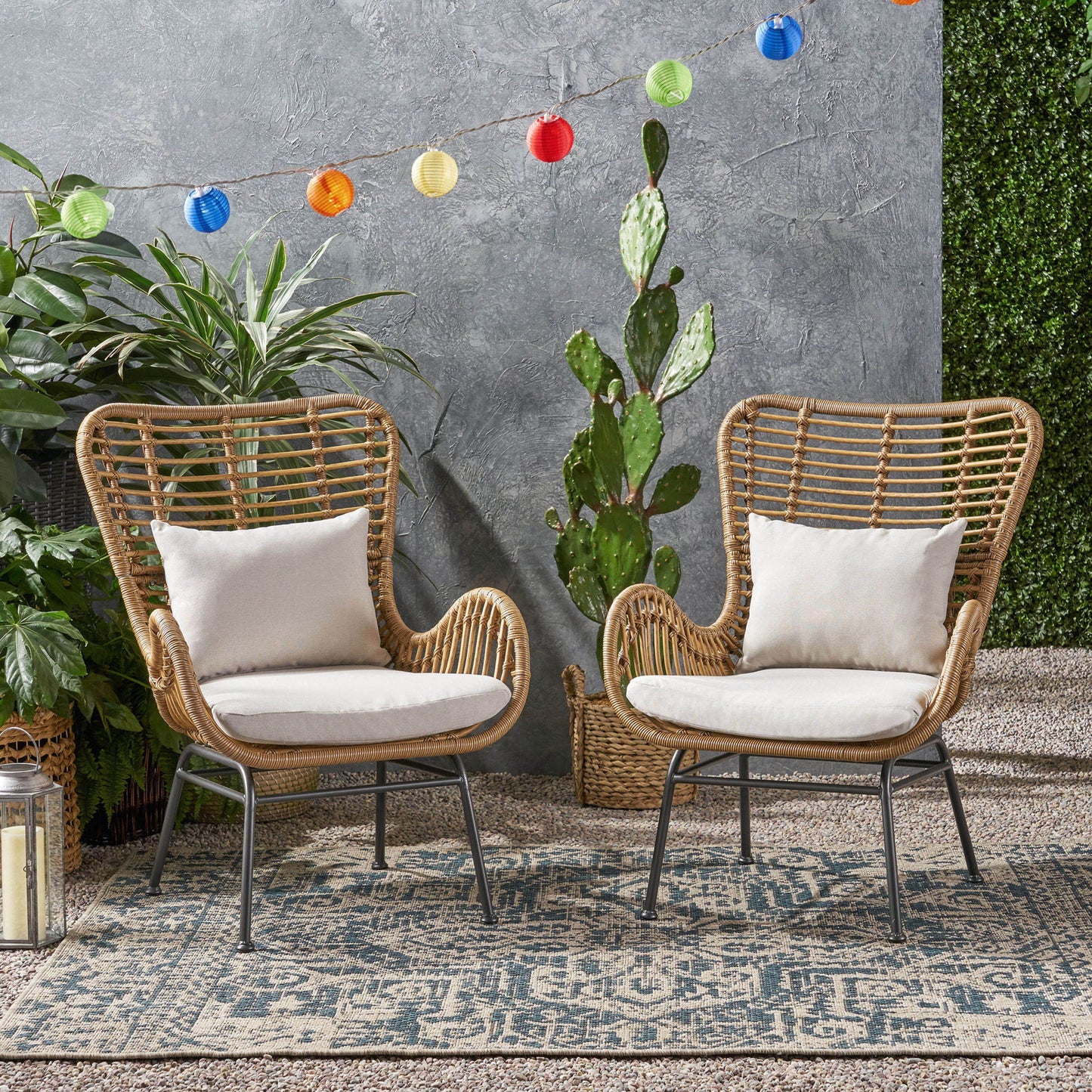 Two Modern Aesthetic Outdoor Rattan Accent Chairs with white cushions sit on a patterned rug among green plants and a cactus, while colorful string lights hang above against a gray textured wall.