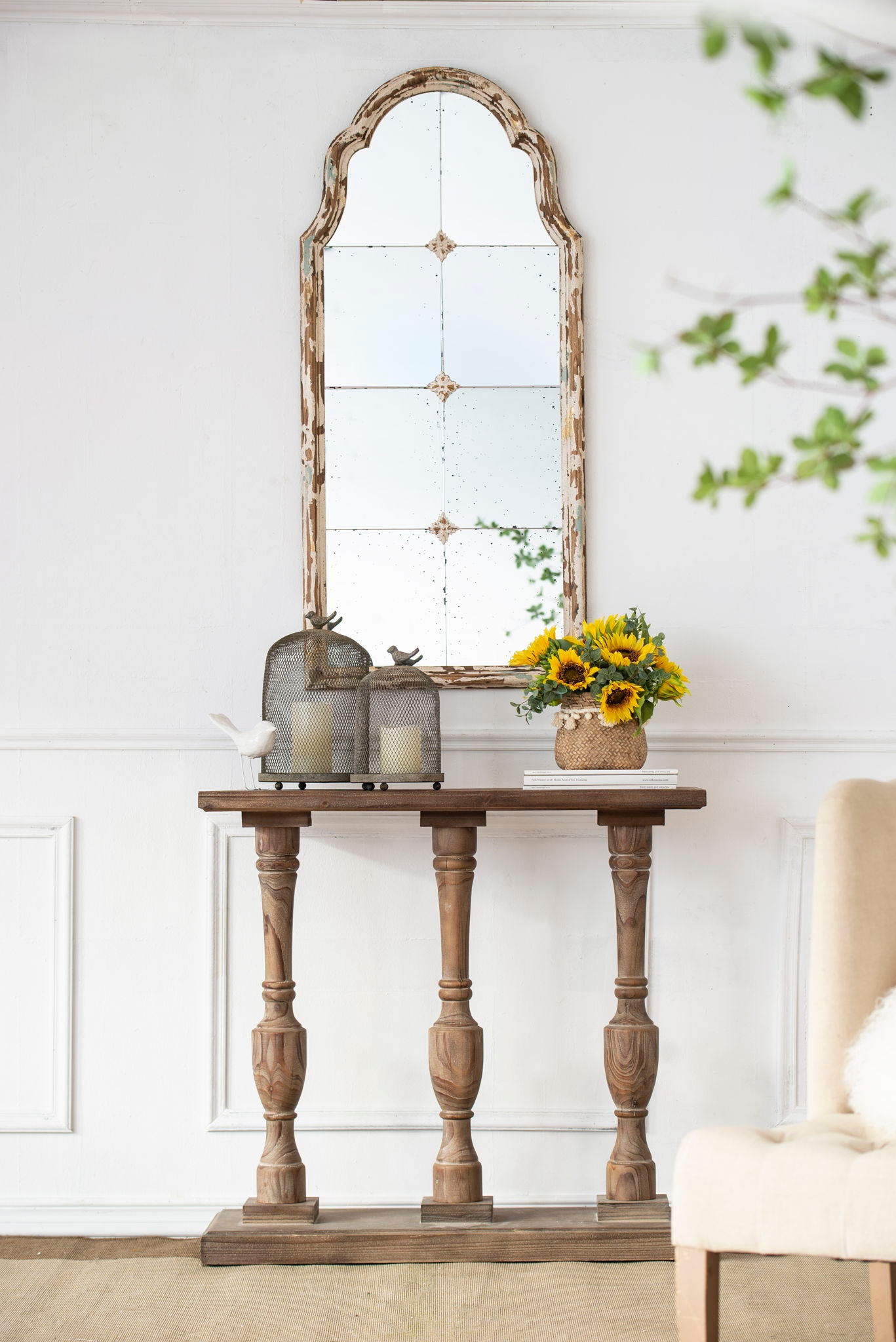 A rustic wooden console table holds two lanterns, a bird figure, and sunflowers. Above, the Large & Framed Wall Mirror—an arched mirror with a decorative window look in cream—hangs on the white wall.