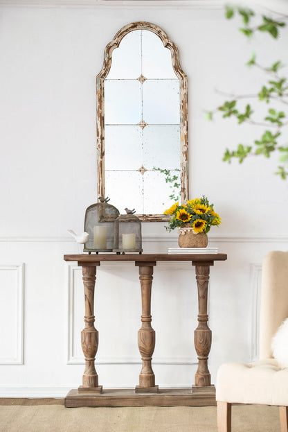 A rustic wooden console table holds two lanterns, a bird figure, and sunflowers. Above, the Large & Framed Wall Mirror—an arched mirror with a decorative window look in cream—hangs on the white wall.