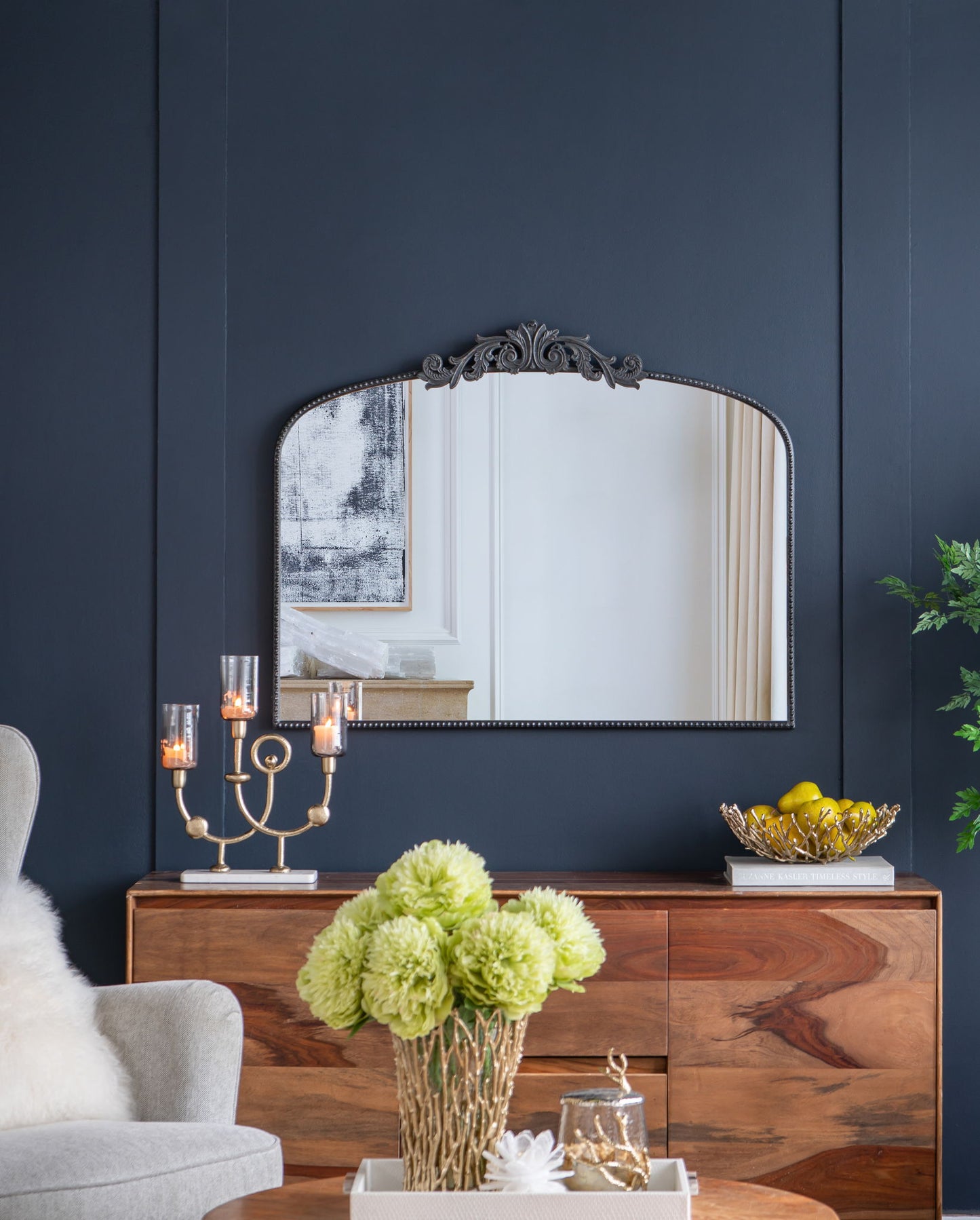 A dark blue wall displays an Arch Mirror, Baroque Inspired Wall Decor above a wooden sideboard holding a candelabra with candles, a glass bowl of lemons, and a green floral vase. A gray armchair appears partially on the left.