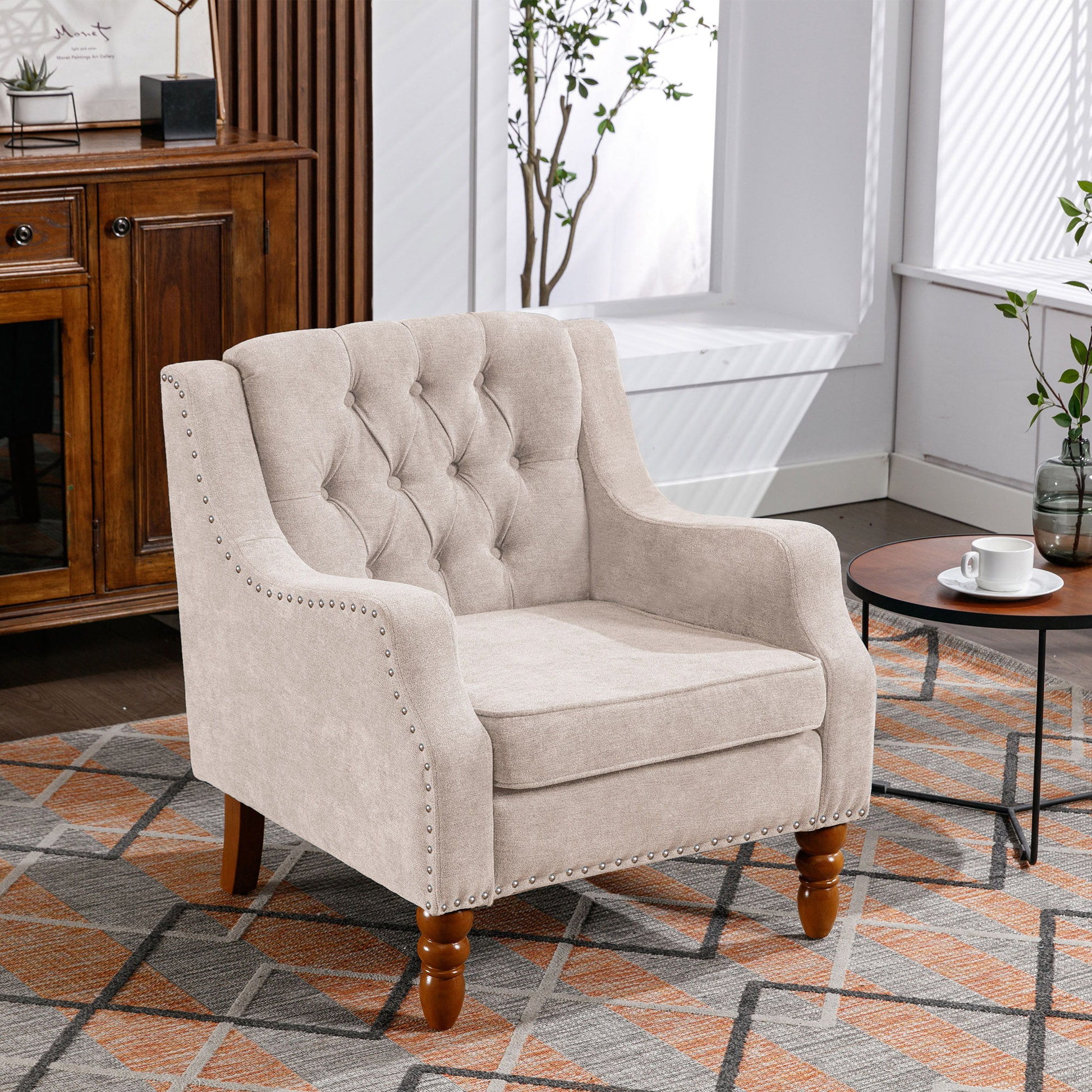 A comfy Accent Chair set with vintage brass studs and button-tufted upholstery is placed on a patterned rug by a window, near a small round table with a cup and saucer, in a bright modern room with a wooden cabinet.