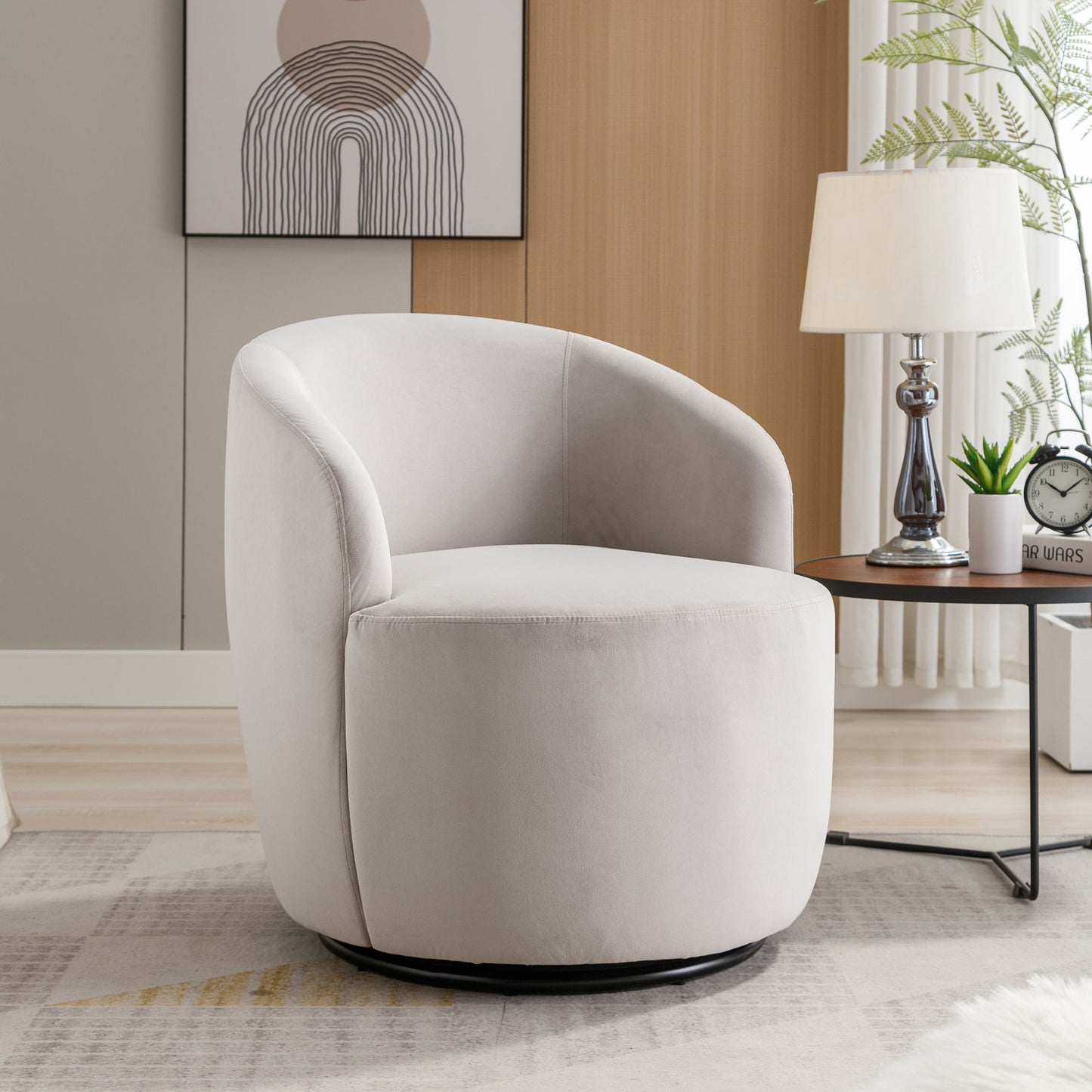 A Velvet Fabric Swivel Accent Armchair Barrel Chair with a powder-coated metal ring sits by a round wooden side table holding a lamp, clock, plant, and books. A minimalist abstract art piece hangs on the wall behind the chair.