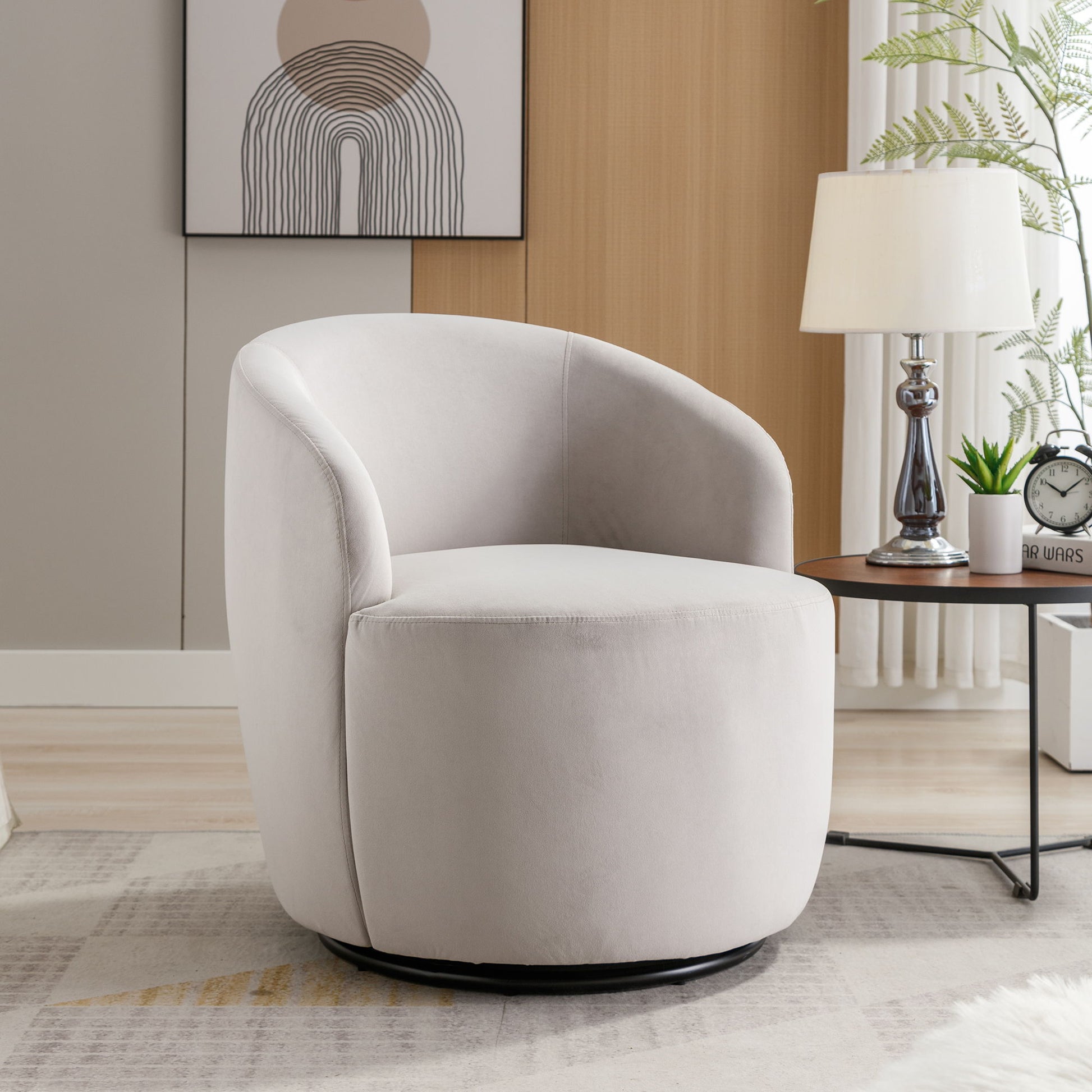 A Velvet Fabric Swivel Accent Armchair Barrel Chair with a powder-coated metal ring sits by a round wooden side table holding a lamp, clock, plant, and books. A minimalist abstract art piece hangs on the wall behind the chair.
