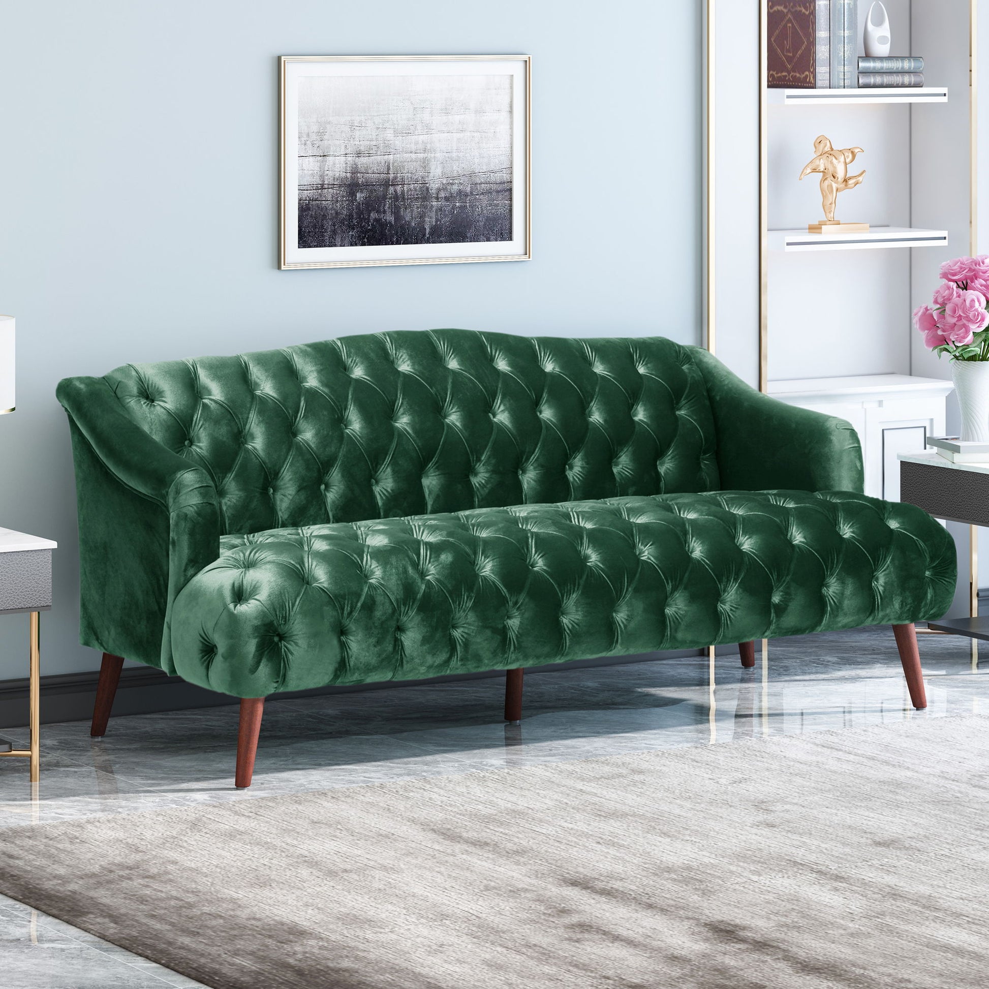 A modern living room with the Comfy 3 Seat Sofa With Wooden Legs in green velvet, a gray rug, wall-mounted abstract art, and a white bookshelf displaying decor and books.