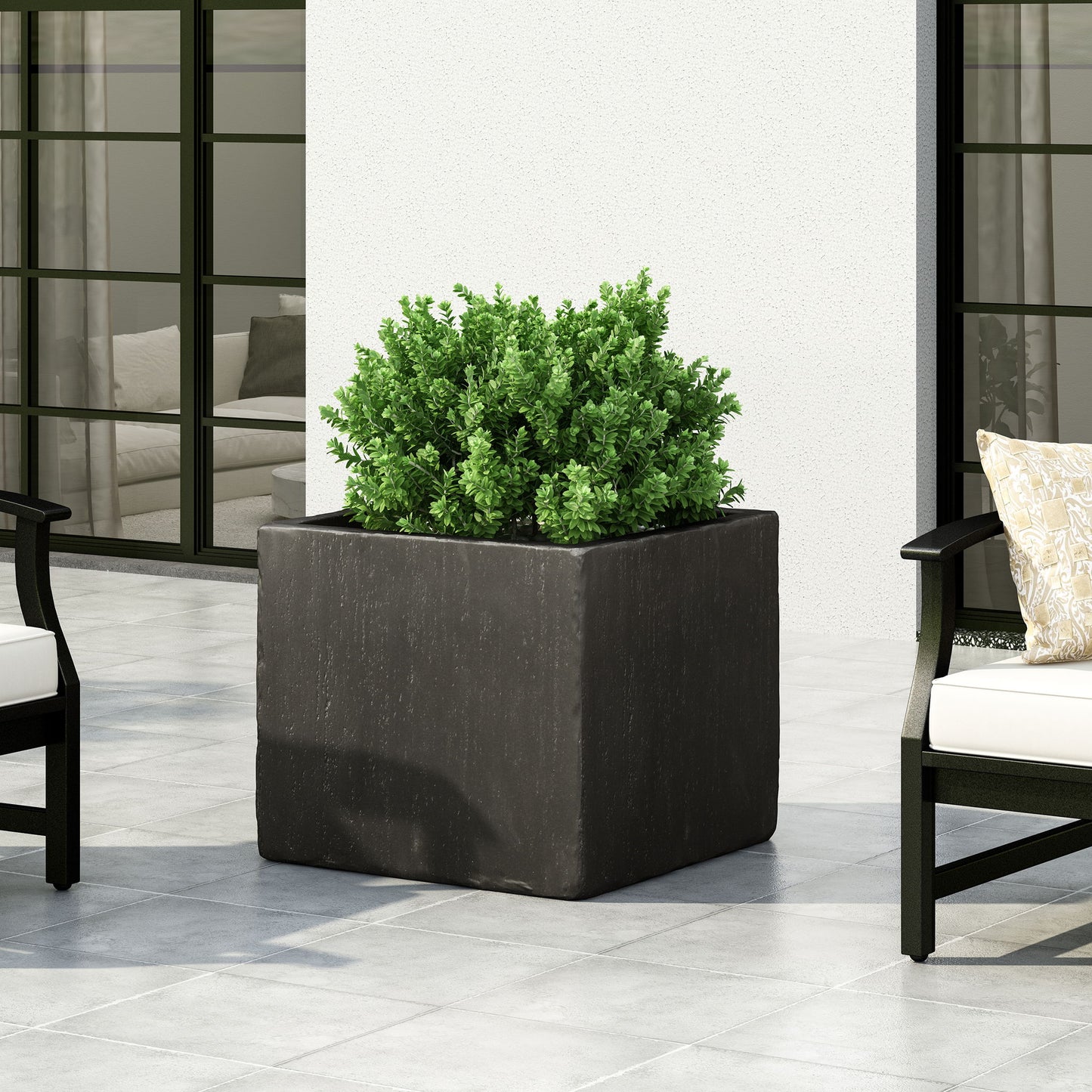 A green shrub thrives in a Modern Outdoor Planter, a large square black container set on a tiled patio, accompanied by modern outdoor chairs with light cushions. Glass doors and a white wall complete the stylish background.