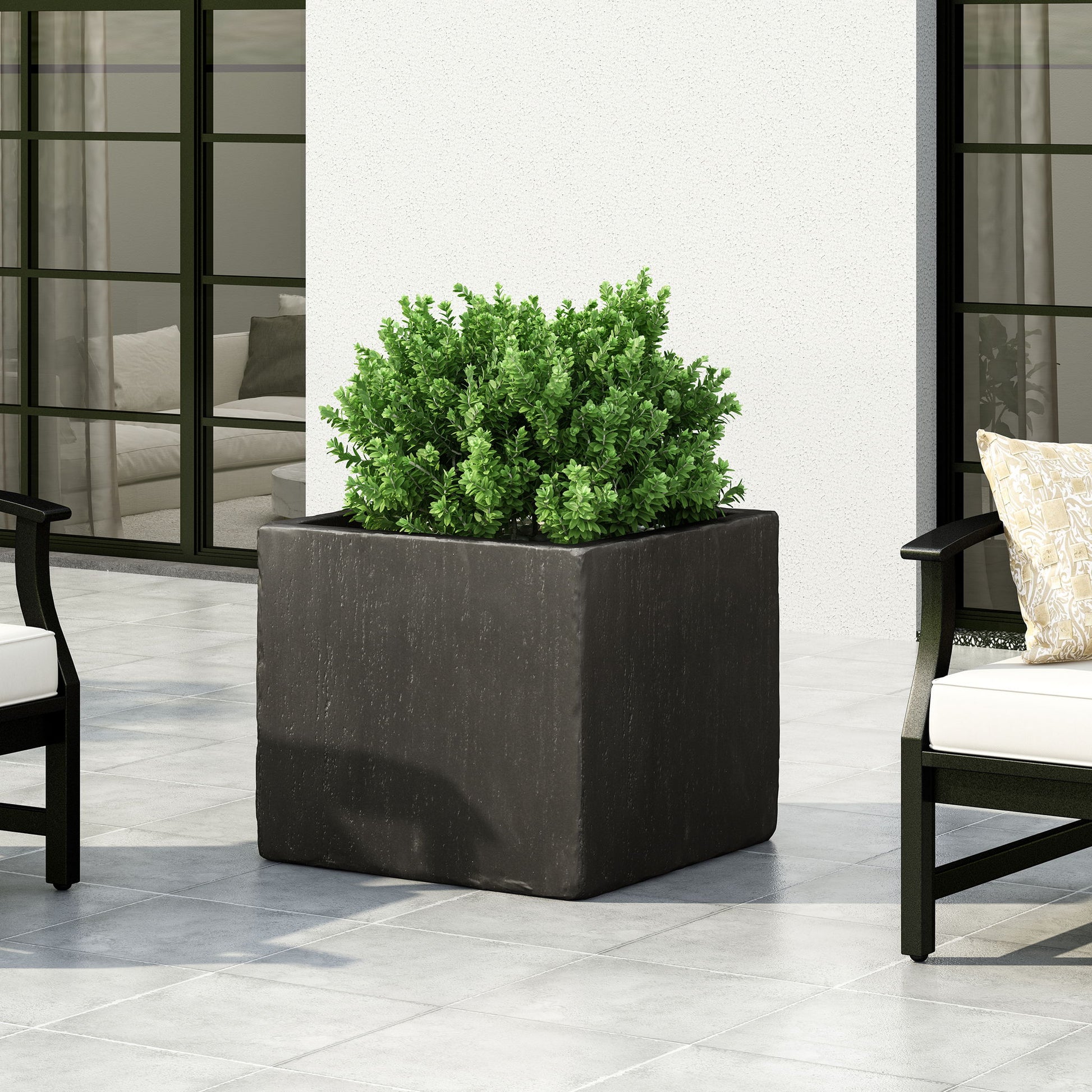 A green shrub thrives in a Modern Outdoor Planter, a large square black container set on a tiled patio, accompanied by modern outdoor chairs with light cushions. Glass doors and a white wall complete the stylish background.