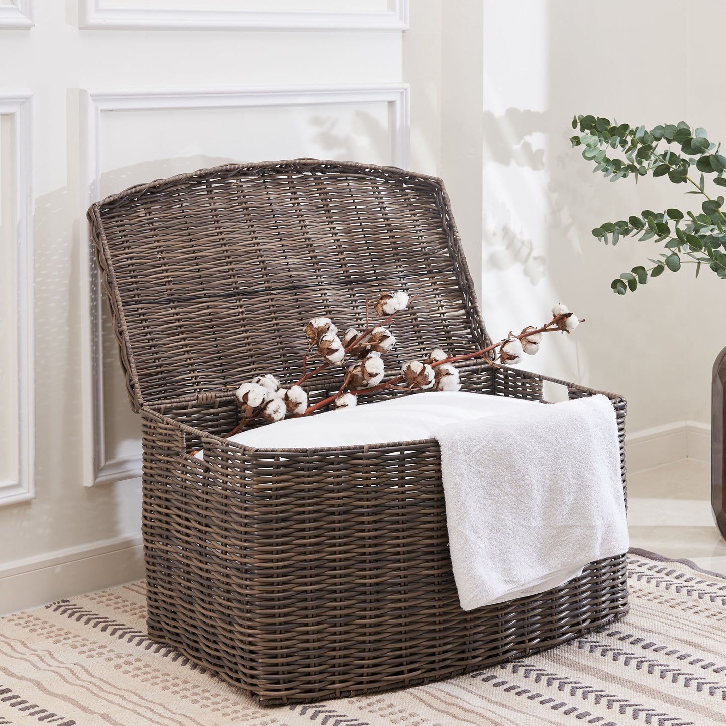 The Joseph Rectangular Curve Resin Woven Wicker Trunk with Handles in Walnut Brown holds a white towel and cotton branch. It sits on a patterned rug beside a wall with white paneling and a vase of green foliage.