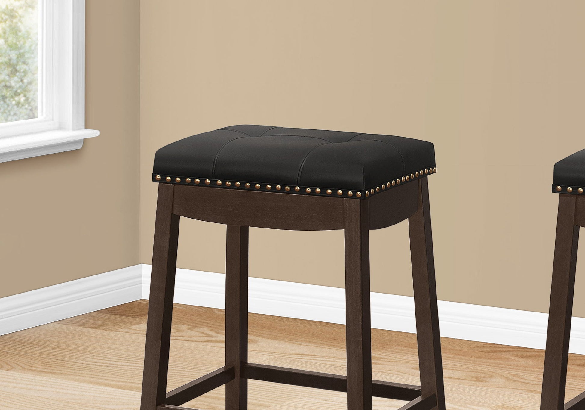 A pair of Bar Stool Counter Height, Saddle Seat, Transitional stools with black cushioned seats and brass stud detailing sit on a light wood floor next to a beige wall and a window with white trim.
