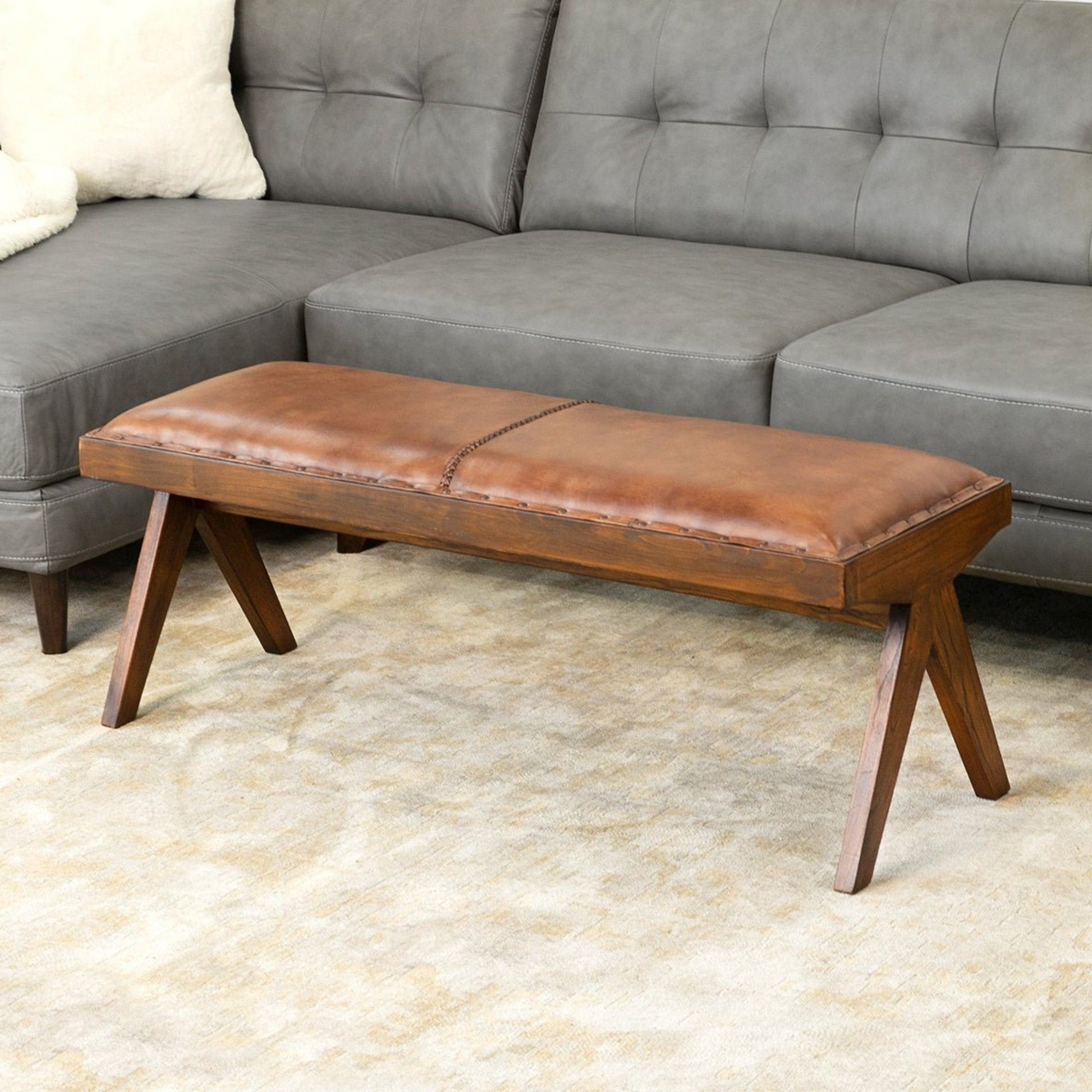 The Chad Mid-Century Modern Leather Bench in brown/tan with cushioned seating stands on a light beige rug before a gray tufted sectional sofa.