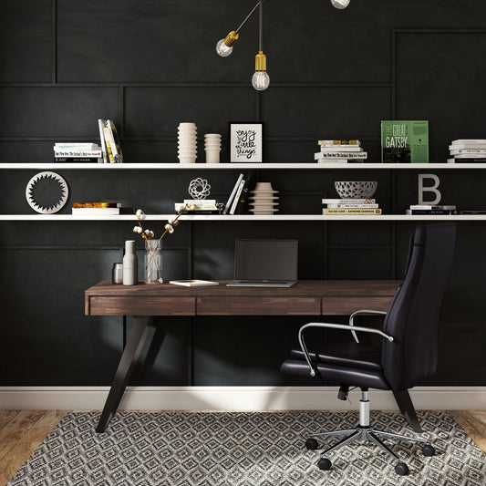 Modern home office with a dark accent wall, floating shelves holding books and decor, the Lowry - Handcrafted Desk with a closed laptop and vase of flowers, plus a black ergonomic chair on a patterned rug.