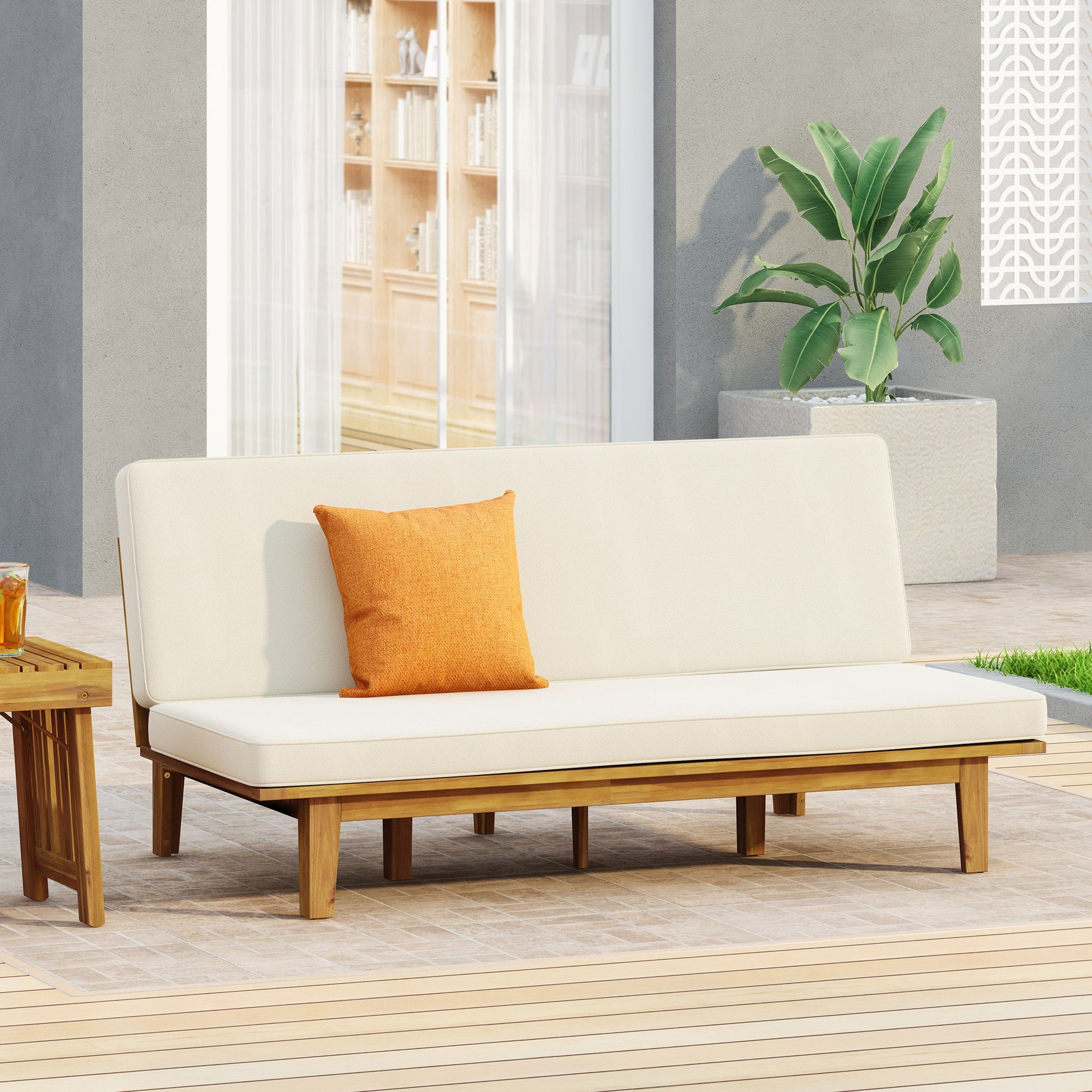 The Outdoor Elegance Daybed With Convertible Design Cushion Frame - Beige is set on a patio, featuring soft beige cushions and an orange pillow beside a small drink table, with a large potted green plant and glass doors in the background.