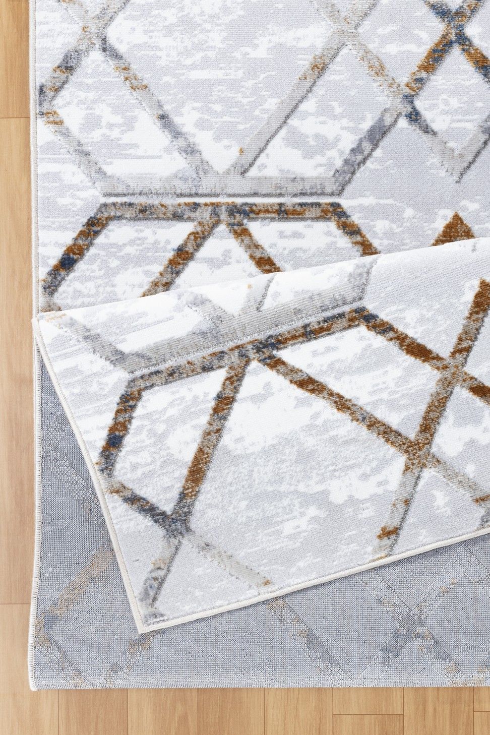 A close-up of the Nova - GC_SOH9001 Area Rug with a geometric pattern in gray, white, and brown on a light wood floor. One corner is folded over to reveal the underside.