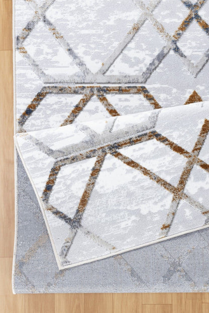 A close-up of the Nova - GC_SOH9001 Area Rug with a geometric pattern in gray, white, and brown on a light wood floor. One corner is folded over to reveal the underside.