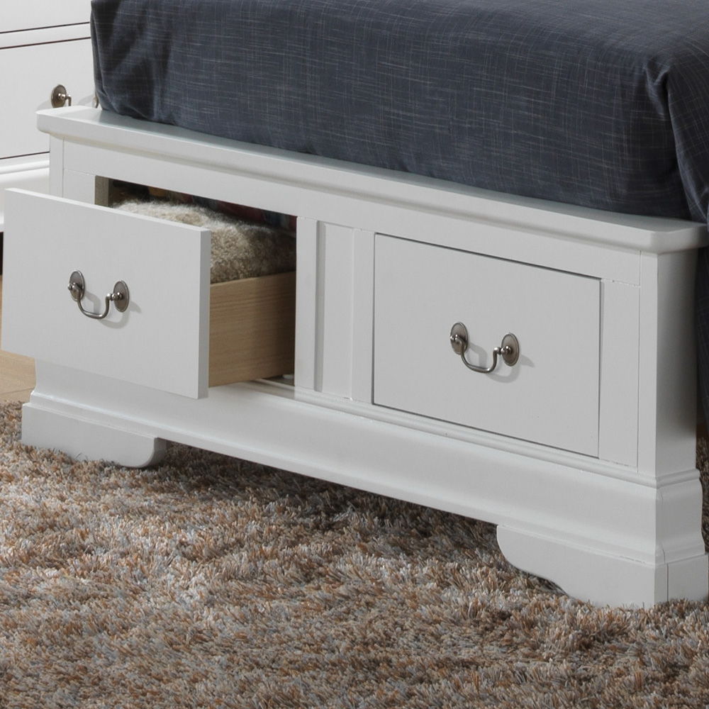 The Louis Phillipe - Storage Bed is a white wooden frame with built-in storage drawers at the foot, one shown open. It's topped with a dark gray blanket and rests on a textured beige and brown carpet.