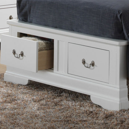 The Louis Phillipe - Storage Bed is a white wooden frame with built-in storage drawers at the foot, one shown open. It's topped with a dark gray blanket and rests on a textured beige and brown carpet.