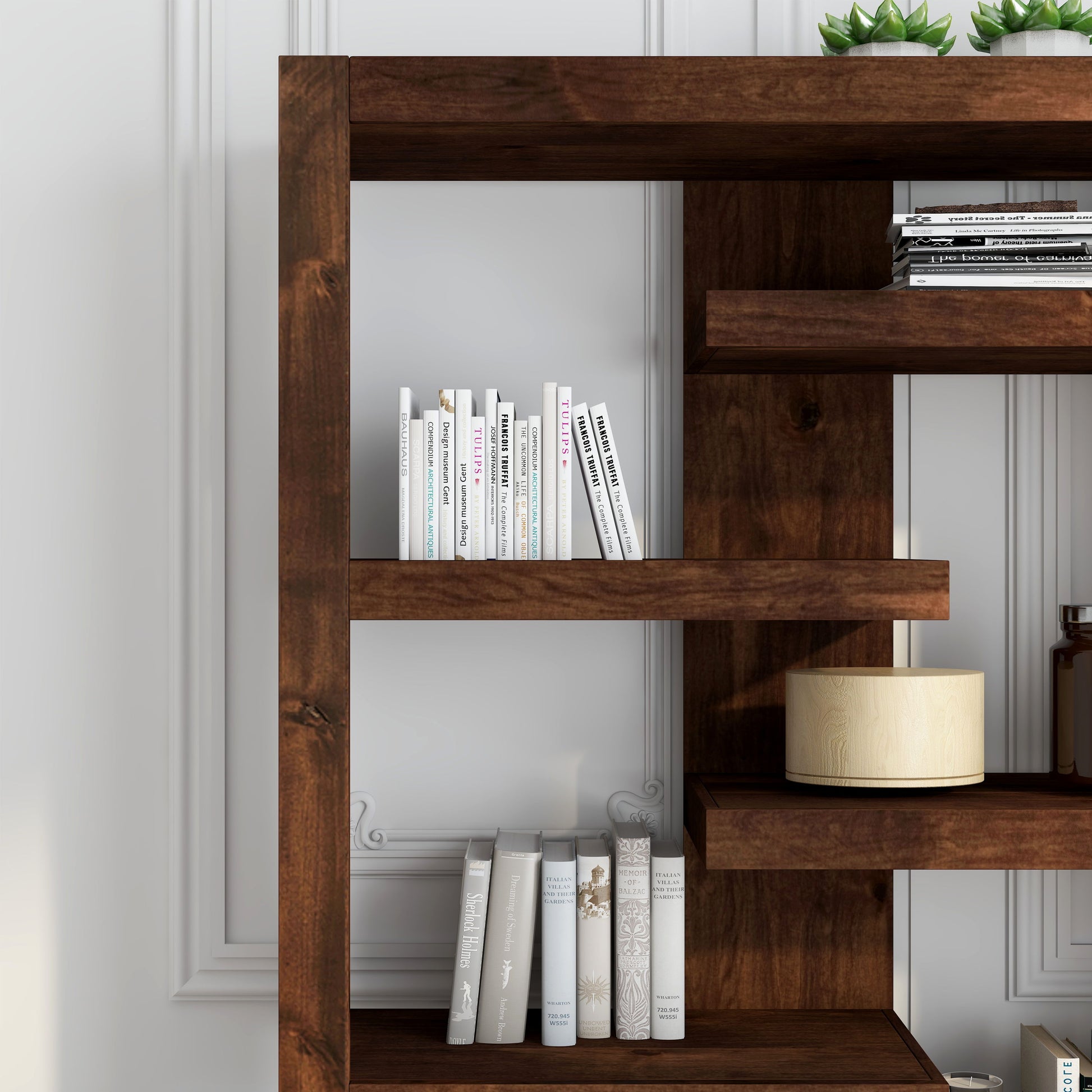 The Couch.com Sausalito 6 Shelf Bookcase in dark brown wood stands against a white paneled wall, displaying neatly arranged books, two small potted succulents, a round beige lamp, and decorative jars.