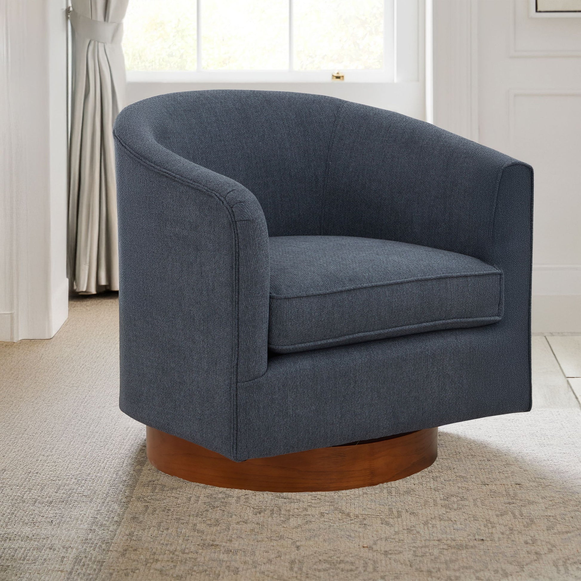 The Miramar Swivel Barrel Chair with a wooden base in dark gray features a modern curved back and sits on a beige patterned rug in a bright room with white walls and a large window.