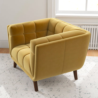The Addison Mid-Century Modern Lounge Chair, in mustard yellow with tufted upholstery and wooden legs, sits on a light patterned rug in a bright room featuring a white wall, large window, and visible radiator.