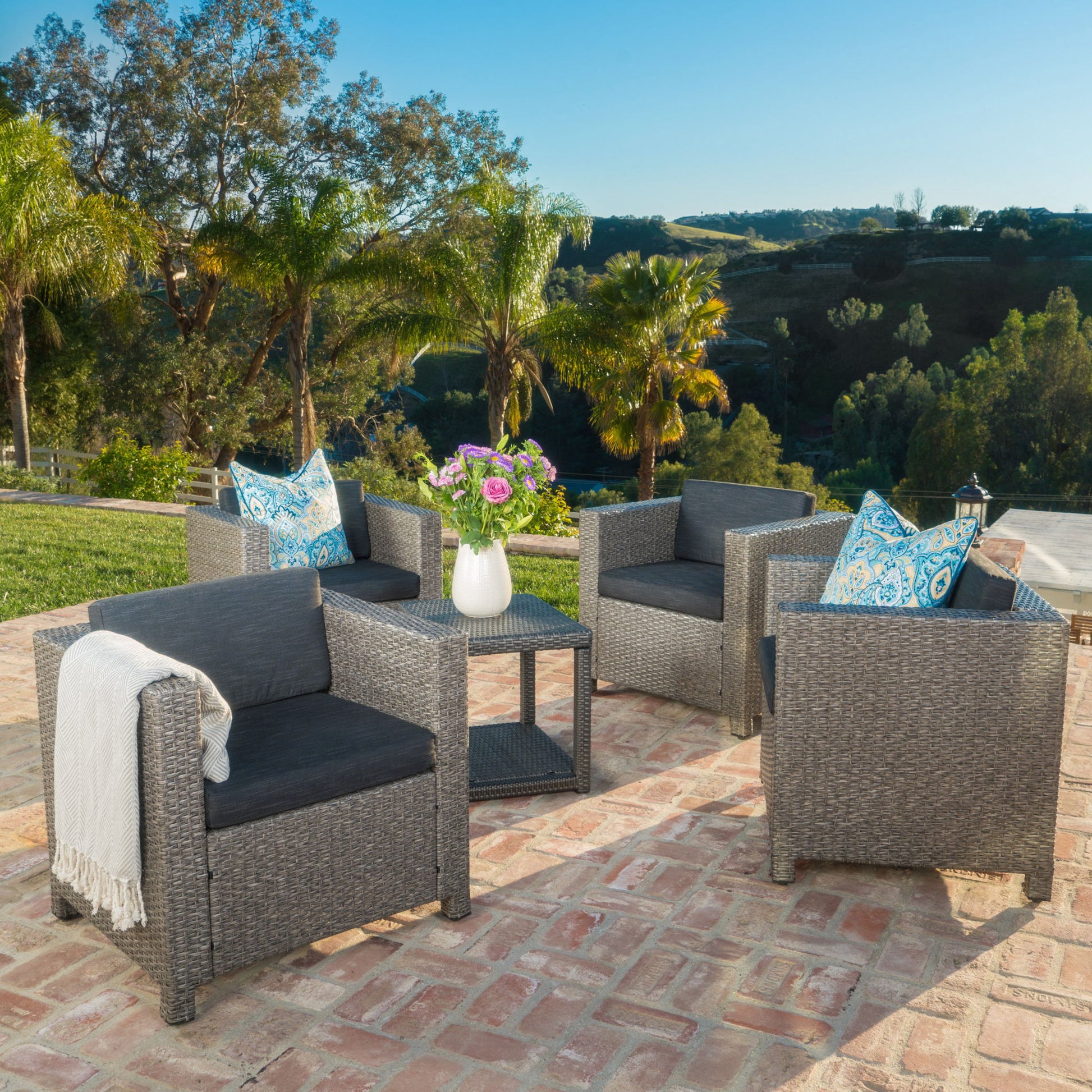 Four Stylish Outdoor Club Chairs with weather resistant cushions surround a small table topped with flowers on a brick patio. Palm trees and greenery are in the background under a clear blue sky.