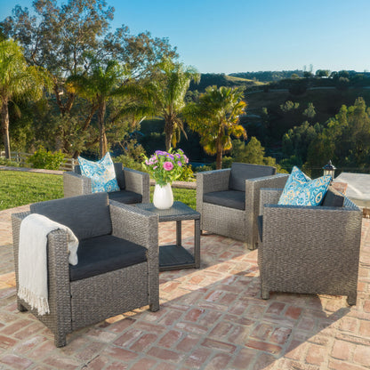 Four Stylish Outdoor Club Chairs with weather resistant cushions surround a small table topped with flowers on a brick patio. Palm trees and greenery are in the background under a clear blue sky.