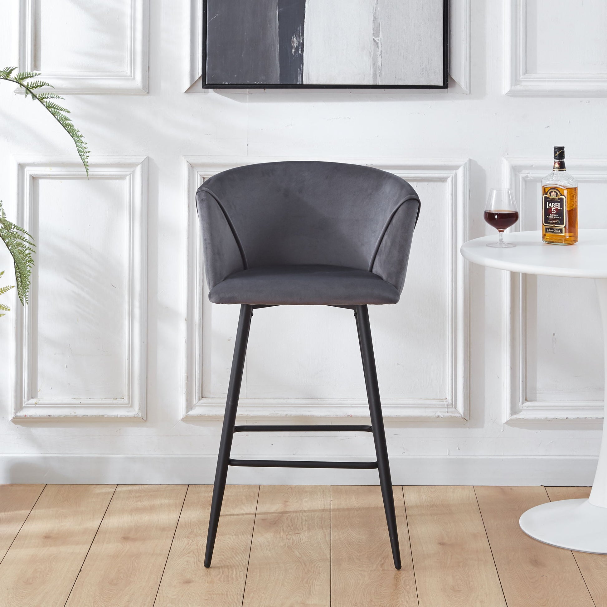 A Counter Height Bar Stool Metal Legs (Set of 2) with curved gray upholstered seats and black metal legs stands on a wooden floor beside a white round table holding a bottle and glass, against a wall with decorative molding.