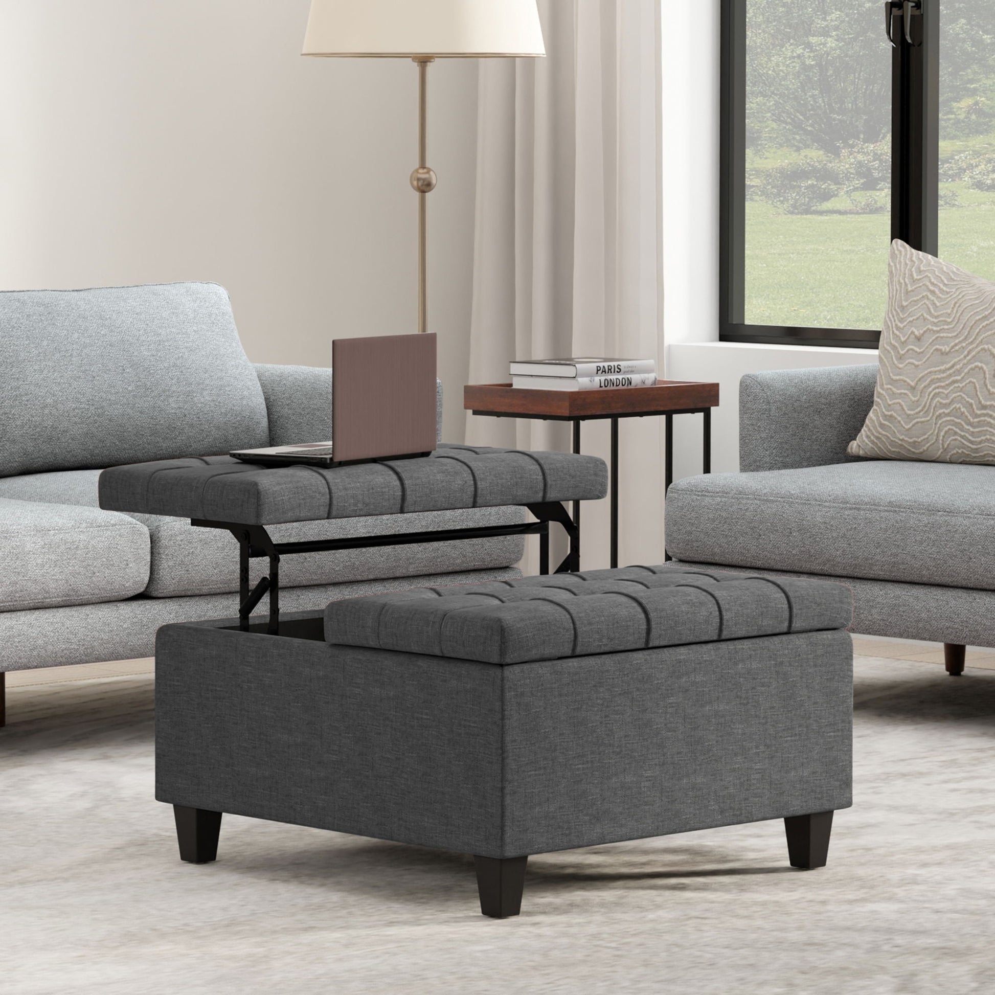 The Harrison Upholstered Coffee Table Storage Ottoman with a lift-top tray sits between two light gray sofas in a modern living room. Nearby, a table with books and a lamp stands beside a large window with white curtains.
