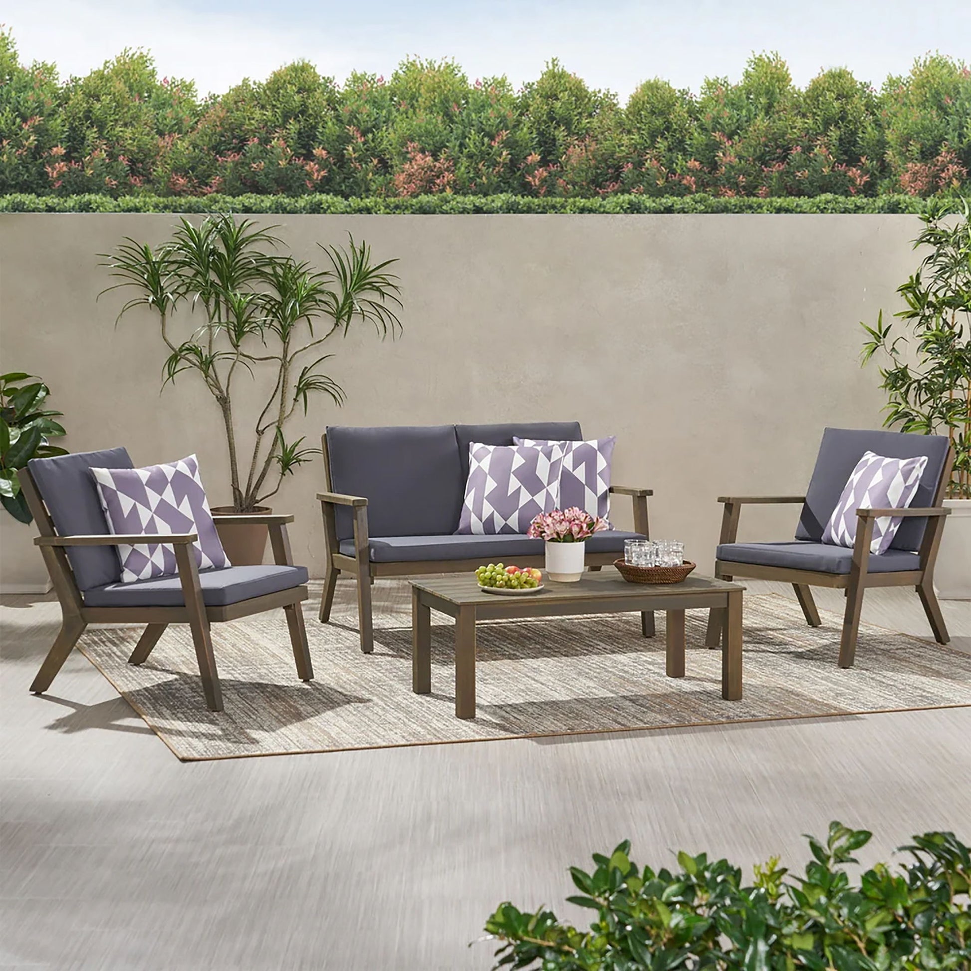 The Avacyn Outdoor Acacia Wood 4 Seater Chat Set with Cushions features a gray cushioned sofa, two chairs with geometric pillows, and a rectangular coffee table on a rug, surrounded by plants, a hedge, and trees.