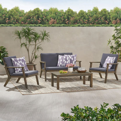The Avacyn Outdoor Acacia Wood 4 Seater Chat Set with Cushions features a gray cushioned sofa, two chairs with geometric pillows, and a rectangular coffee table on a rug, surrounded by plants, a hedge, and trees.