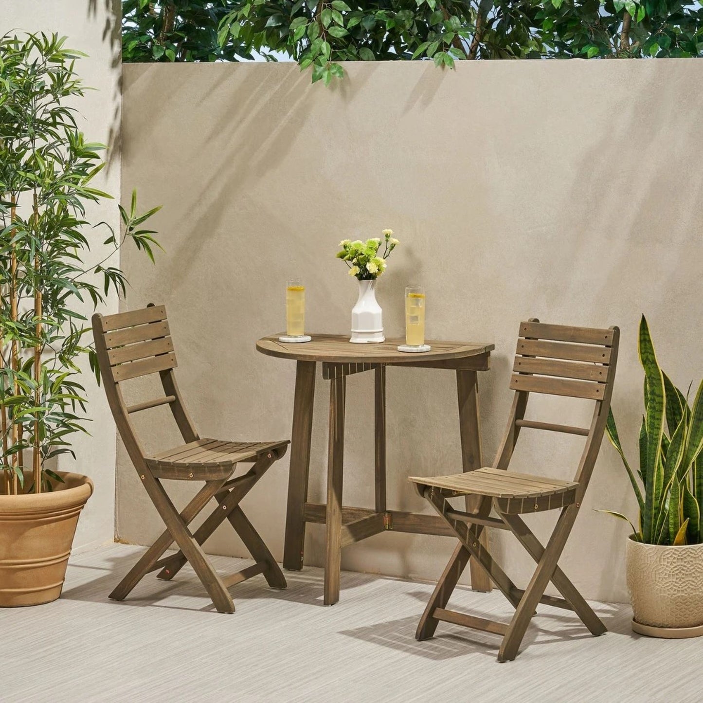 The Outdoor 3 Piece Half Round Folding Bistro Set features two wooden chairs and a round table, styled with two glasses of lemonade, a white vase with green flowers, surrounded by potted plants and a beige wall accented with greenery.