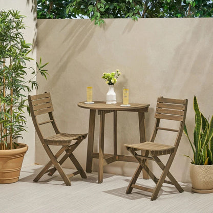 The Outdoor 3 Piece Half Round Folding Bistro Set features two wooden chairs and a round table, styled with two glasses of lemonade, a white vase with green flowers, surrounded by potted plants and a beige wall accented with greenery.