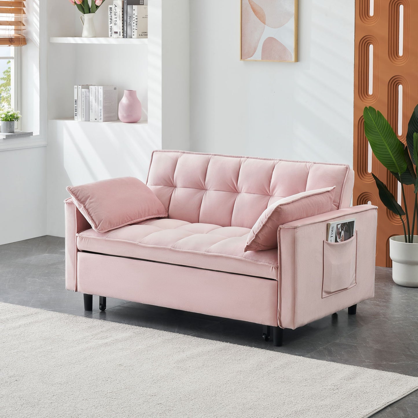 A modern living room features the Modern Velvet Sofa, a 3-in-1 convertible pull-out bed loveseat with back, matching pillows, and side pockets. The space has white walls, a large rug, bookshelves, plants, and abstract framed art.