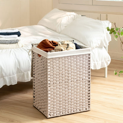 A Laundry Hamper With Lid PE Rattan and powder coating frame, featuring two removable bags, sits on a wooden floor next to a neatly made bed with white bedding. Folded clothes are stacked on the bed in the bright, tidy bedroom.