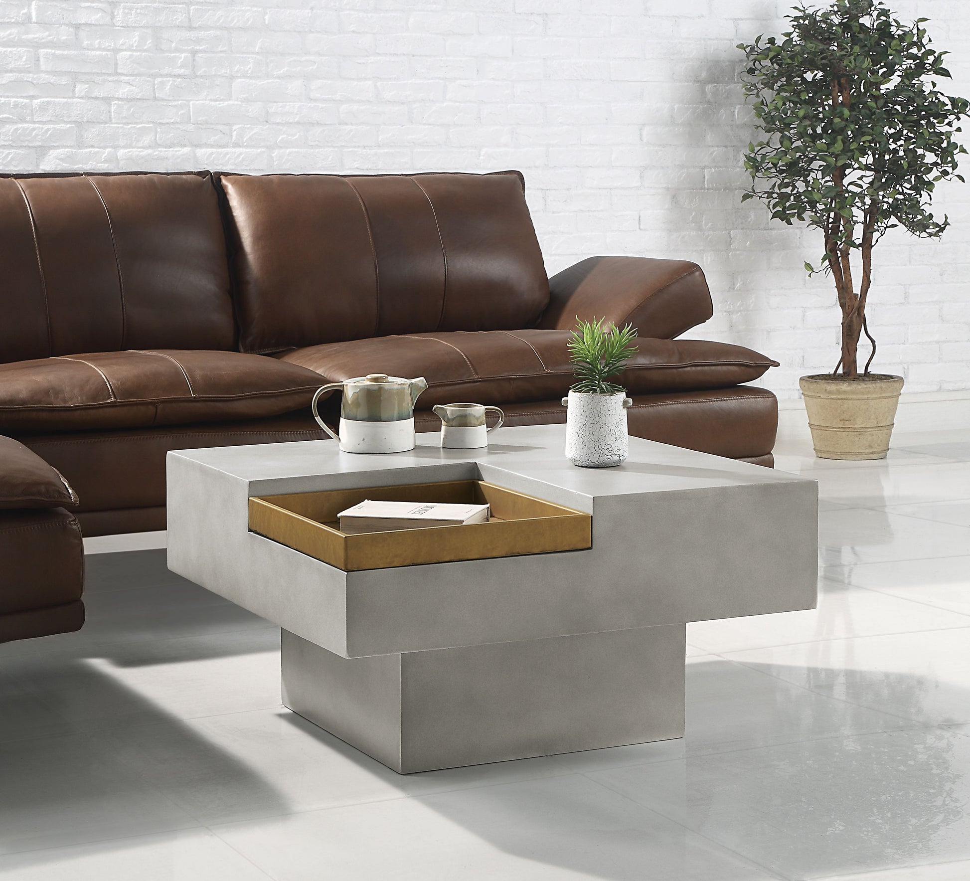 A modern living room with a brown leather sofa, the Kailano Weathered Table in gray featuring an open storage compartment and a potted plant on top, plus a larger potted plant on the floor against a white brick wall.