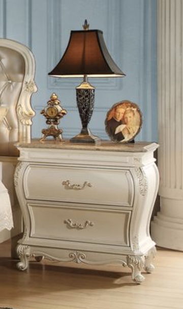 A Chantelle nightstand with a granite top in white, featuring ornate handles and two drawers, supports an intricate table lamp, vintage clock, and framed picture. The setup complements a room adorned with elegant decor and wooden flooring.