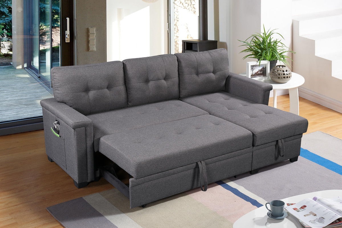 The Ashlyn Reversible Sleeper Sectional Sofa with Storage Chaise, USB Charging Ports, and Pocket is unfolded in a bright living room with wooden floors, a side table plant, stairs, and large windows letting in natural light.