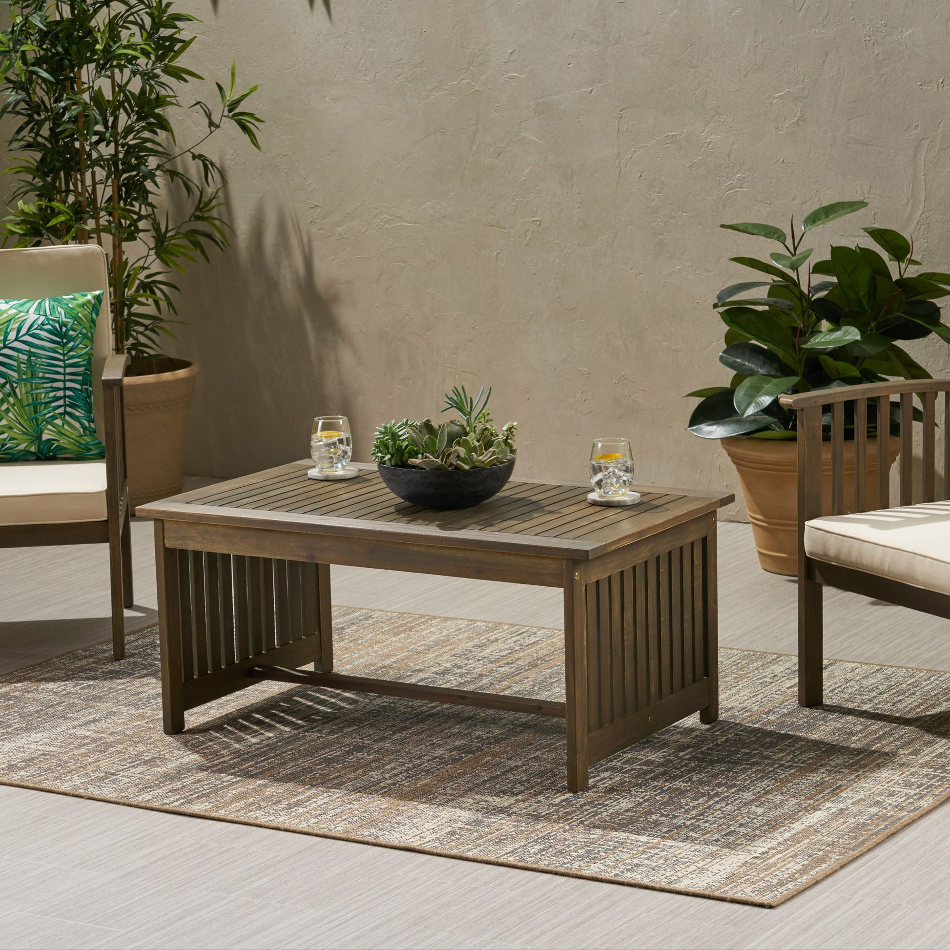 A wooden outdoor seating area features the Acacia Wood Rectangular Patio Coffee Table For Outside, topped with a bowl of succulents and two glasses of water, surrounded by potted plants, cushioned chairs, and a neutral textured rug on the patio.