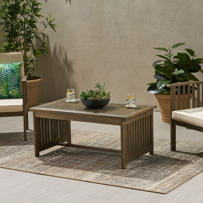 A wooden outdoor seating area features the Acacia Wood Rectangular Patio Coffee Table For Outside, topped with a bowl of succulents and two glasses of water, surrounded by potted plants, cushioned chairs, and a neutral textured rug on the patio.