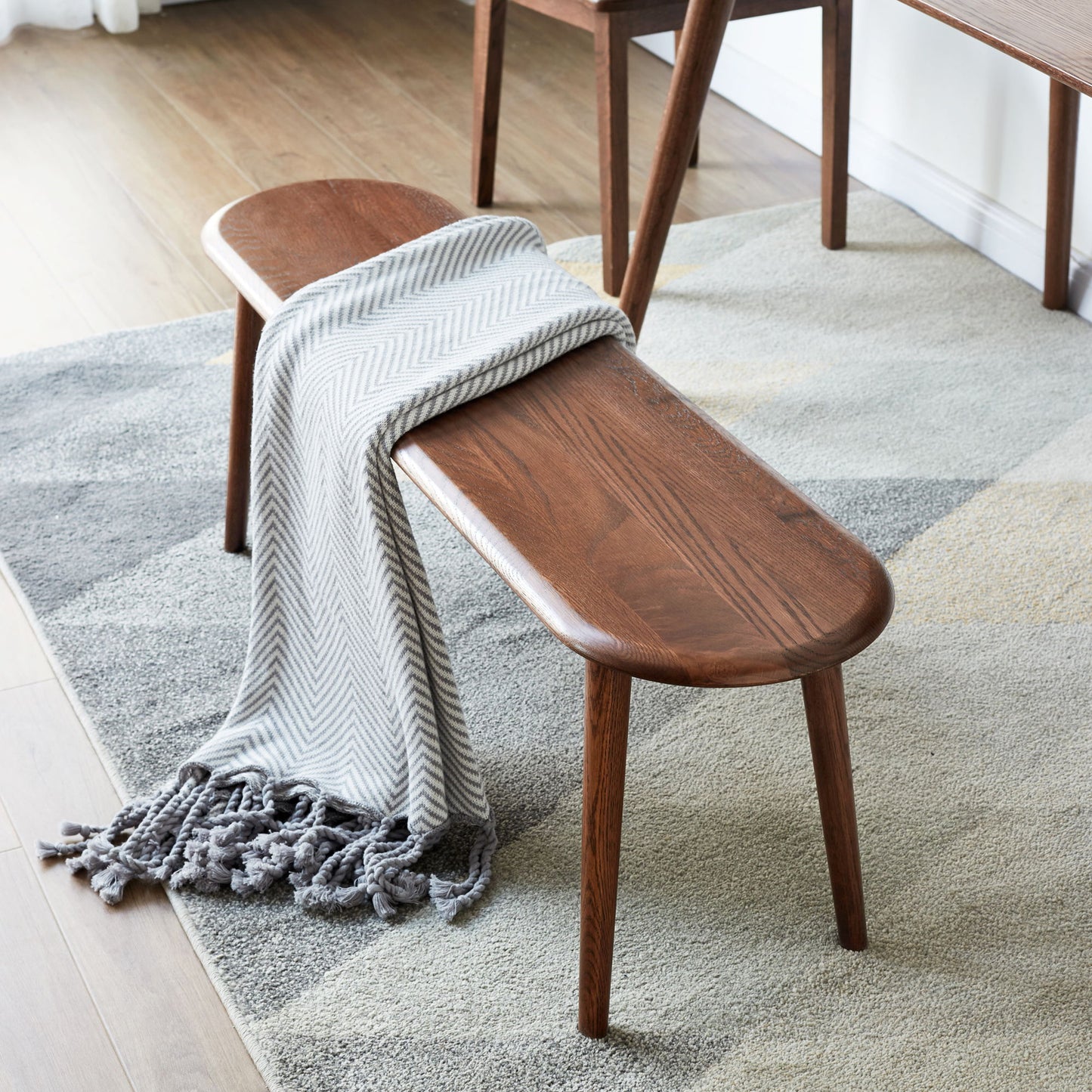 A Dining Bench, Table Bench For Living Room with tapered legs rests on a geometric rug. A gray and white herringbone throw with tassels drapes over one end, while warm light highlights the wood floor in the background.