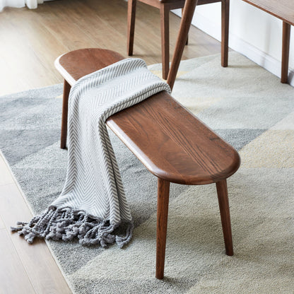 A Dining Bench, Table Bench For Living Room with tapered legs rests on a geometric rug. A gray and white herringbone throw with tassels drapes over one end, while warm light highlights the wood floor in the background.