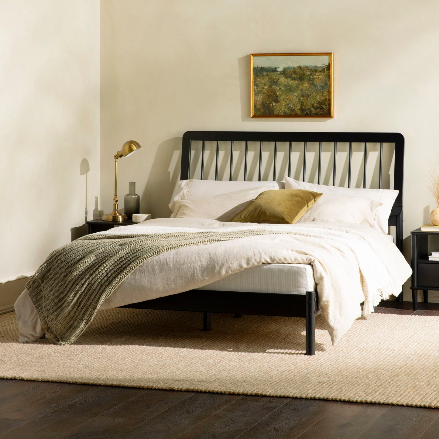 A modern bedroom features the Mid-Century Modern Solid Wood Spindle Bed, styled with beige bedding, a green throw blanket, and a gold accent pillow. Above hangs a framed landscape painting; nightstands and lamps flank each side.