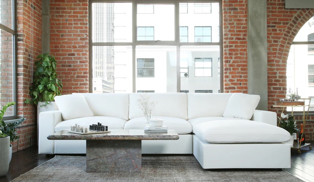 A spacious, modern living room with a large white sectional sofa against a brick wall. A low rectangular coffee table in front of the sofa displays a chess set and vase. Tall windows allow natural light to flood in, highlighting industrial and contemporary elements.