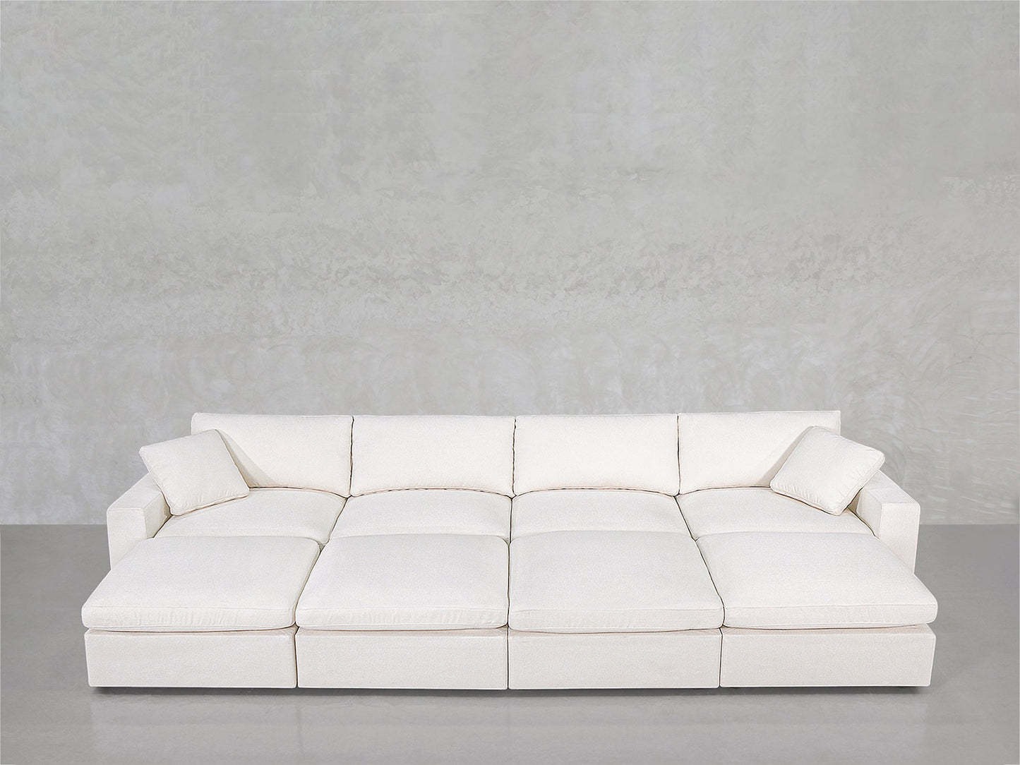 The 8-Seat Modular Daybed is a modern white sectional with wide seats and two matching pillows, shown against a light gray textured wall and smooth gray floor.