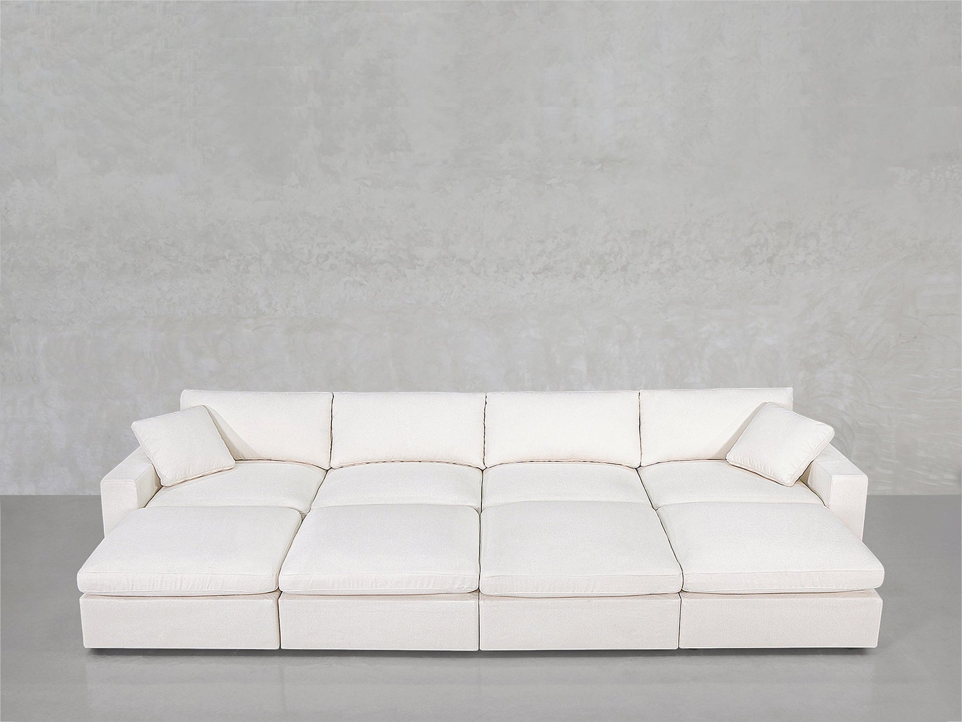 The 8-Seat Modular Daybed is a modern white sectional with wide seats and two matching pillows, shown against a light gray textured wall and smooth gray floor.