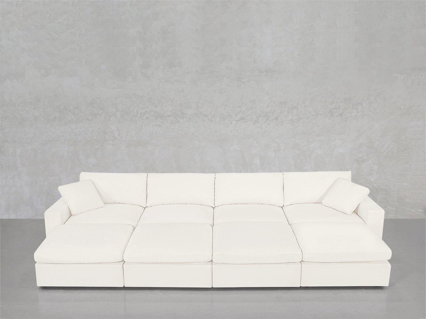 The 8-Seat Modular Daybed, a modern white sectional with wide seats and four back cushions, sits against a light gray wall and gray floor. Two matching square pillows are placed at each end of the sofa.